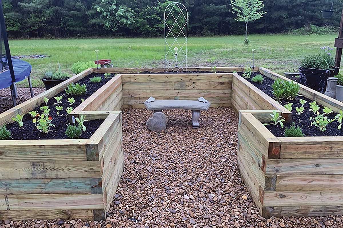 U-shaped wooden raised garden bed surrounding a stone bench, set on a gravel pad in a backyard.